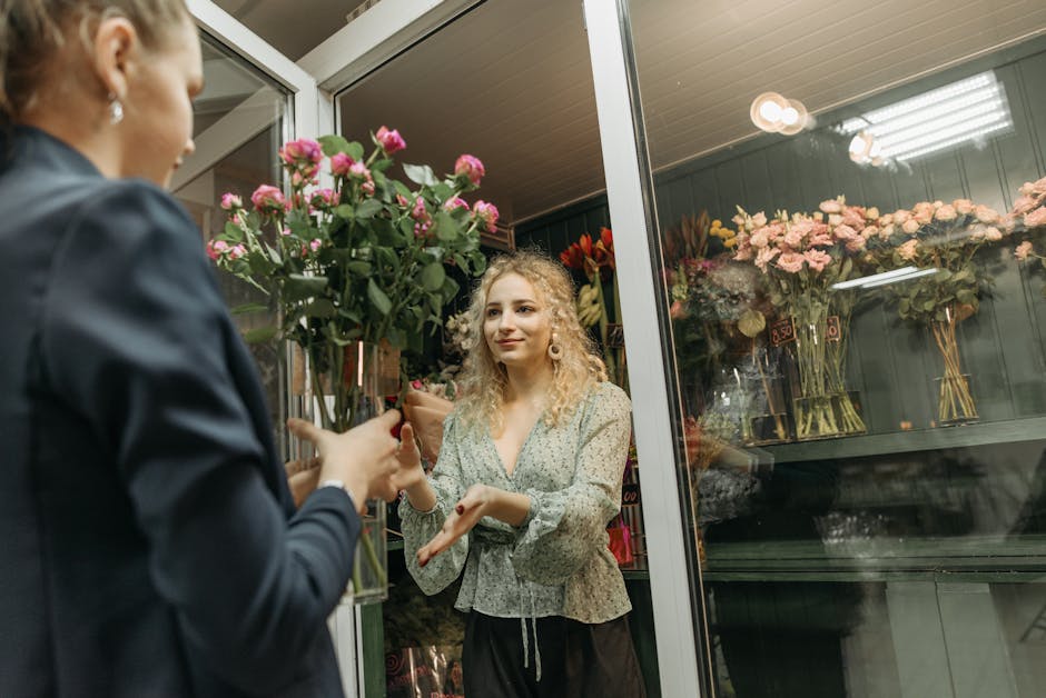 A woman with curly blonde hair dressed in a floral blouse and black trousers is inside a flower shop, receiving a bouquet of pink roses wrapped in brown paper from a florist. The florist, dressed in a