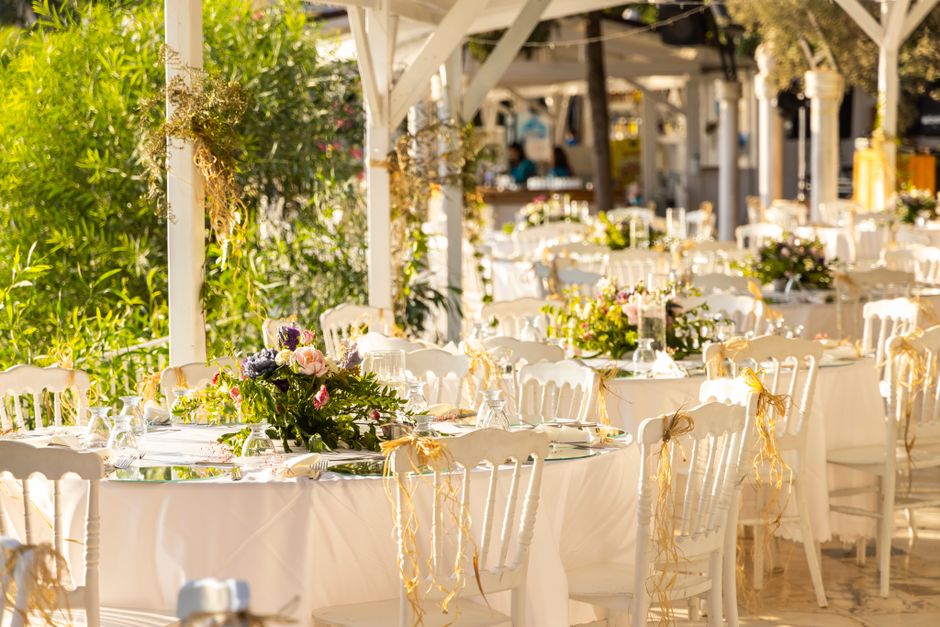 An outdoor event space decorated with elegant floral arrangements on white round tables covered with white tablecloths. The floral centerpieces feature a mix of lush pink, purple, and white flowers, i