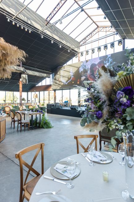 An elegantly decorated event space with a high, glass-paneled ceiling and modern industrial design. In the foreground, a round table is set with white tablecloths, clear glassware, silver cutlery, and