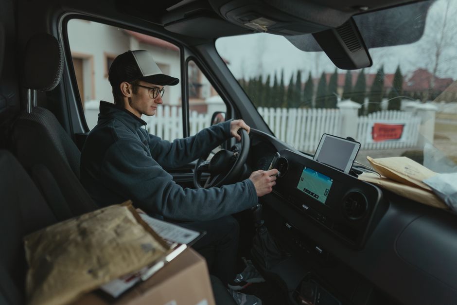 A delivery driver seated in the front of a van, preparing to deliver a floral gift. The interior of the van shows a modern dashboard with a digital display, and the driver is holding the steering whee