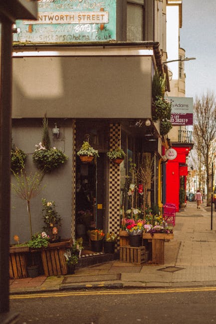 The image depicts the exterior of a florist shop on a street, with several flower arrangements and potted plants displayed outside. The flowers include vibrant pink, yellow, and white blossoms arrange