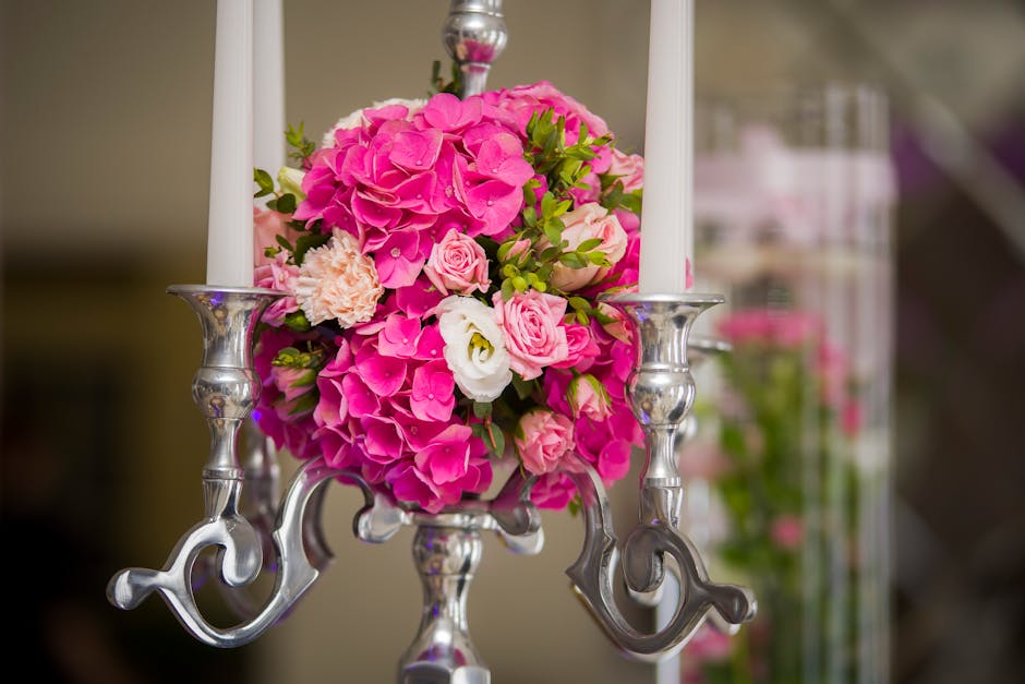 A floral arrangement featuring a bouquet of vibrant pink hydrangeas, light pink roses, and creamy white lisianthus, complemented by green foliage, placed in a silver vase and arranged on a reflective 