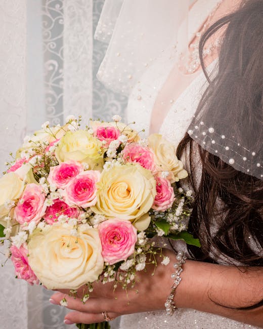 A close-up of a bride holding a floral bouquet with a mix of cream-colored roses and pink spray roses, accented by small white baby's breath flowers and lush green foliage. The bouquet is wrapped in w